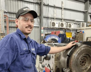 A photo of Gavin wearing a cap and eye protection glasses with his left hand touching a machine. - Ryder Machining Specialists
